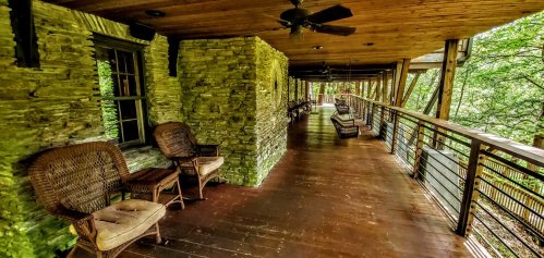 A spacious porch with wooden flooring, stone walls, and wicker chairs, surrounded by lush greenery.