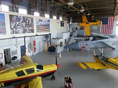 A spacious hangar with various aircraft, including a yellow biplane and a sleek gray jet, and an American flag on the wall.