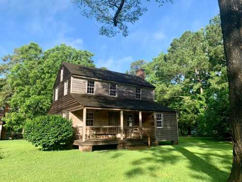 A two-story wooden house with a porch, surrounded by lush green trees and grass under a clear blue sky.