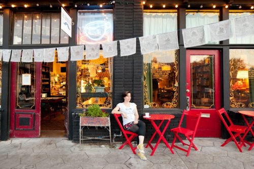A woman sits at a red table outside a cozy café, decorated with string lights and colorful banners.