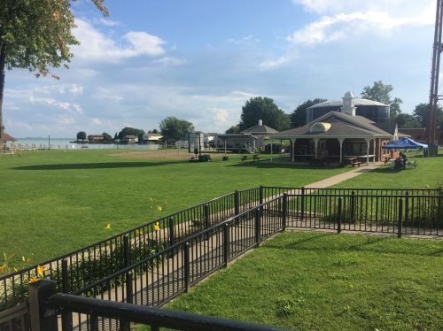 A grassy area with a gazebo and picnic tables, overlooking a lake under a blue sky with scattered clouds.
