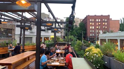 A rooftop restaurant with diners enjoying meals amidst greenery and city buildings at dusk.