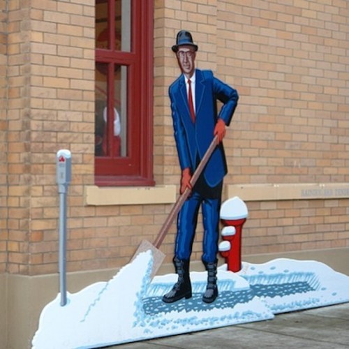 A painted figure in a suit and hat shovels snow beside a fire hydrant, set against a brick wall.