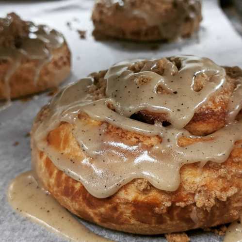 Close-up of a glazed donut topped with crumbly streusel and drizzled with a creamy glaze on a parchment-lined surface.