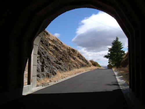 A view through a tunnel arch, revealing a road lined with rocky hills and a blue sky with clouds.