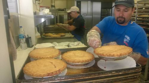 A baker in a blue shirt prepares pies on a tray in a busy kitchen, with another worker in the background.