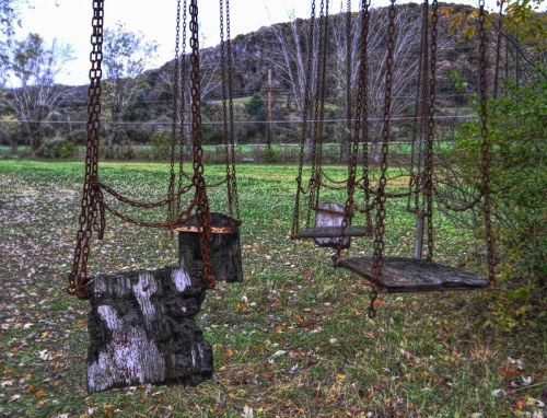 Rusty swings hang from chains in a grassy area, surrounded by trees and a distant hill.