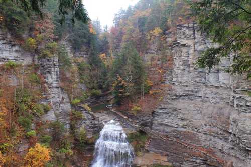 A serene waterfall cascades through rocky cliffs, surrounded by vibrant autumn foliage and lush green trees.