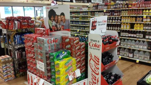 Display of Coca-Cola products and various soft drinks in a grocery store aisle, with promotional signage.