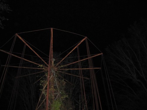 Rusty, abandoned amusement park ride silhouetted against a dark night sky, surrounded by bare trees.