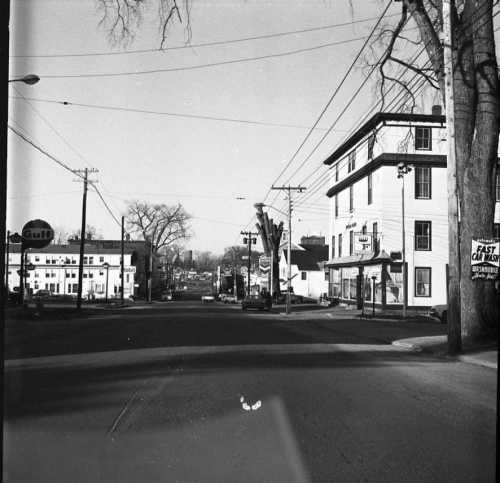 Black and white photo of a street scene with buildings, power lines, and trees on a clear day.