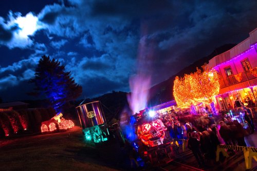A colorful night scene featuring a train with Halloween decorations, surrounded by illuminated trees and a festive crowd.