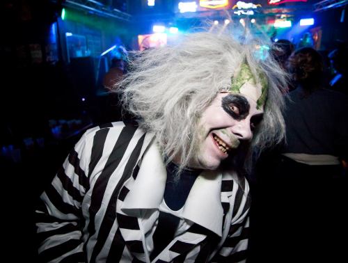 A person in a striped suit and wild hair, with face paint, smiles at a lively party scene.