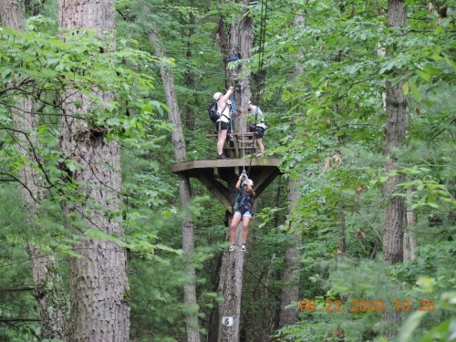 Two people on a zip line platform in a forest, with one person descending on the zip line.