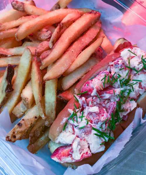 A lobster roll topped with chives, served with a side of golden French fries in a takeout container.