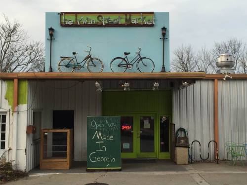 Exterior of a market with bicycles on the roof, a green door, and a sign reading "Open Now Made In Georgia."