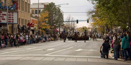 A parade scene with horseback riders and a crowd of spectators along a city street lined with autumn trees.