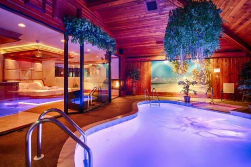 Indoor pool area with a tropical mural, wooden ceiling, and soft blue lighting, featuring plants and a hot tub.