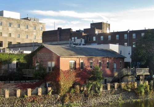A brick building with greenery along a riverbank, surrounded by urban structures under a clear blue sky.