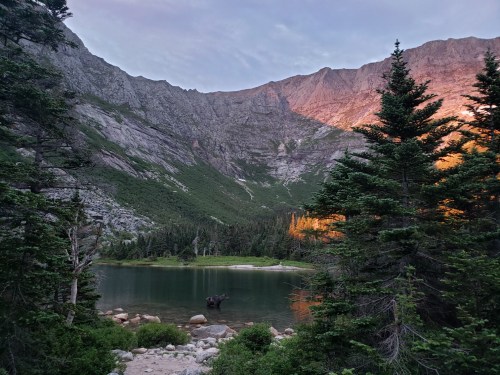 A serene mountain landscape with a calm lake, surrounded by trees and rocky cliffs under a twilight sky.