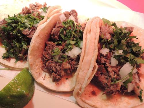 Three tacos filled with shredded meat, topped with chopped onions and cilantro, served with a lime wedge.