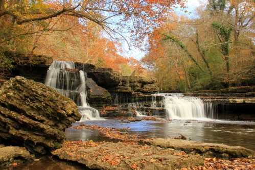 A serene waterfall cascades over rocky ledges, surrounded by autumn foliage and a tranquil river.