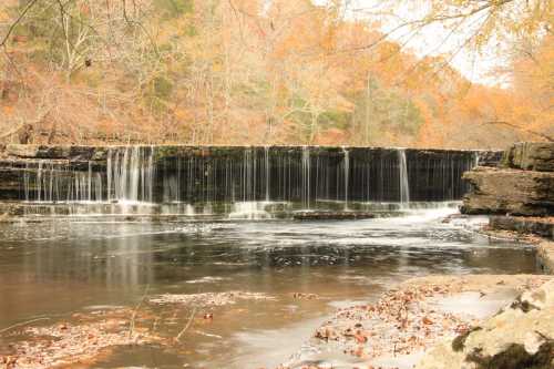 A serene waterfall cascades over a rocky ledge, surrounded by autumn foliage and a calm river.