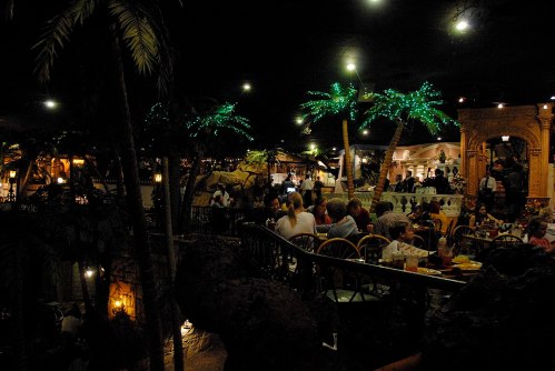 A lively restaurant scene with palm trees, dim lighting, and diners enjoying meals in a tropical-themed setting.