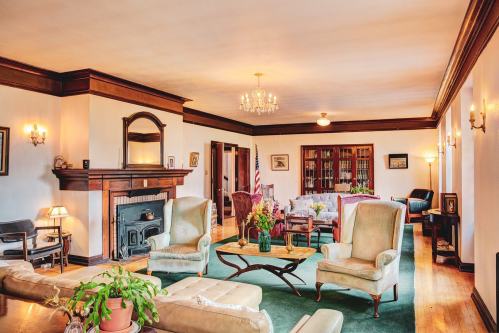 A spacious living room with elegant furniture, a fireplace, and decorative plants, featuring warm lighting and wooden accents.