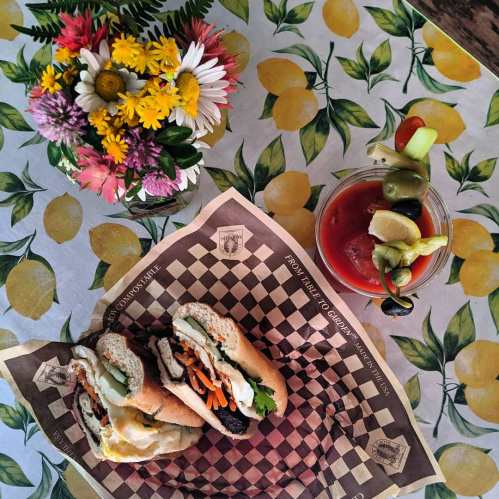 A colorful table setting with two sandwiches, a cocktail, and a vase of flowers on a lemon-patterned tablecloth.