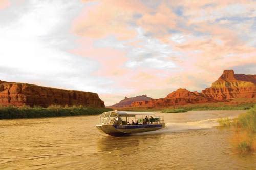 A boat with passengers navigates a river surrounded by red rock formations under a colorful sky.