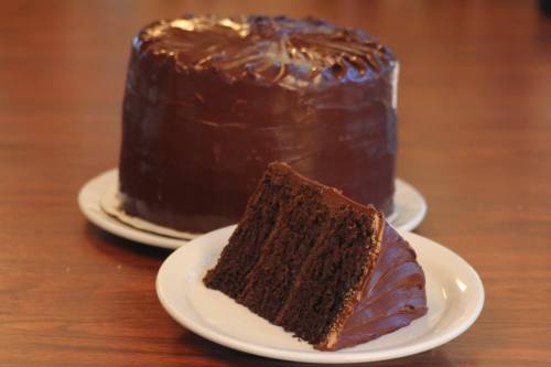 A rich chocolate cake with a slice cut out, displayed on a white plate against a wooden table background.