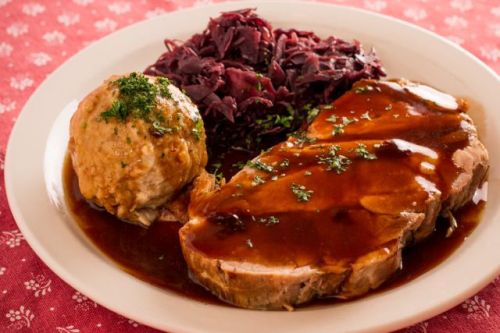 A plate of sliced roast meat with gravy, a dumpling, and red cabbage on a patterned tablecloth.