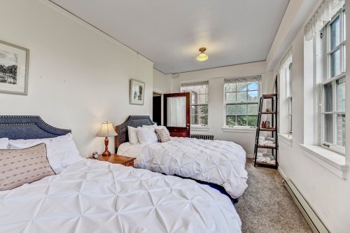 Bright bedroom featuring two beds with white bedding, a nightstand, and large windows letting in natural light.