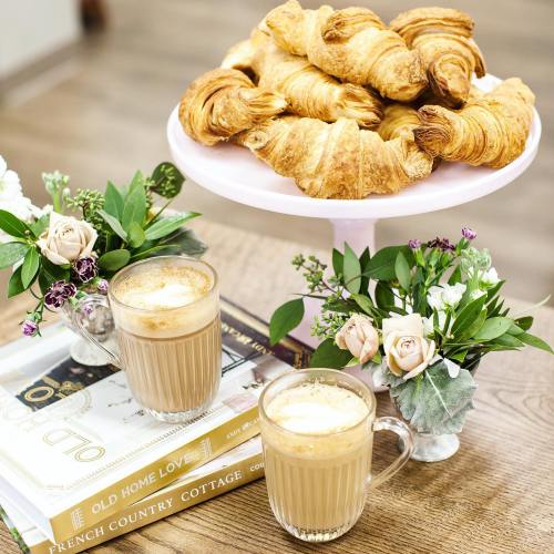 A table with two coffee cups, a stack of croissants on a pink stand, and small floral arrangements.