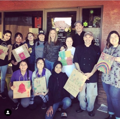A group of diverse individuals poses outside a restaurant, holding decorated paper bags and smiling.