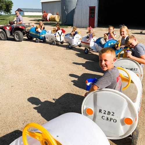 Children ride in colorful toy train cars on a dirt path, while an adult drives a quad nearby.