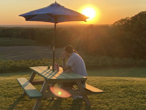 A person sits at a picnic table under an umbrella, watching the sunset over a scenic landscape.