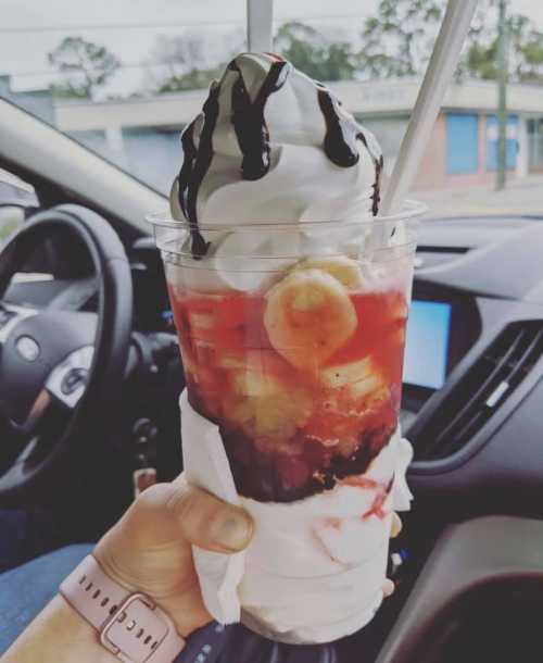 A hand holding a large dessert cup with soft serve ice cream, chocolate drizzle, and fruit, inside a car.