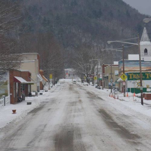 Snow-covered street lined with small shops and trees, set against a backdrop of mountains on a cloudy day.