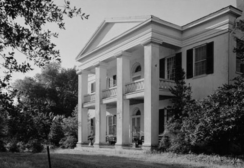 Historic mansion with tall columns, large windows, and a porch, surrounded by trees and greenery. Black and white photo.