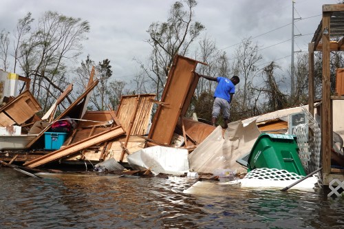 A person navigates debris and floodwaters after a storm, with damaged furniture and structures in the background.