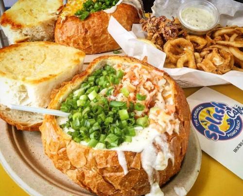 A bowl of creamy soup topped with green onions, served in a bread bowl, alongside onion rings and toasted bread.