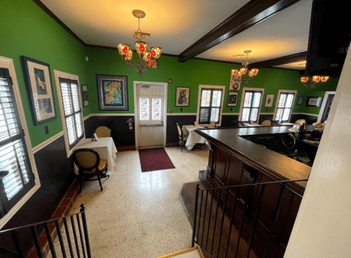 A cozy restaurant interior with green walls, decorative chandeliers, and tables set for dining near a bar area.