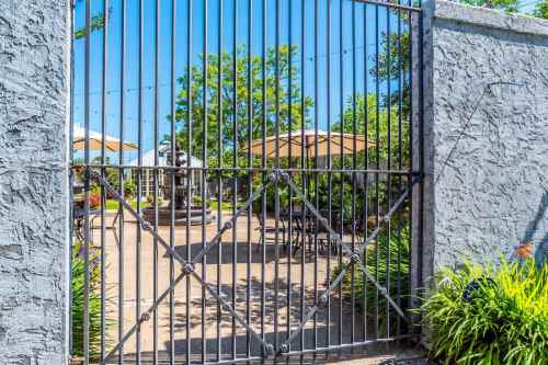 A gated entrance to a sunny outdoor patio with tables, umbrellas, and lush greenery.