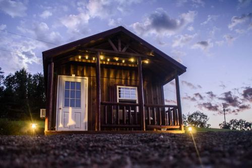 A small wooden cabin with a porch, illuminated by lights, set against a twilight sky with scattered clouds.