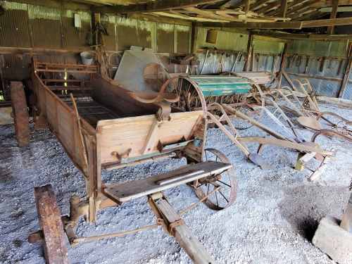 An old barn filled with vintage farming equipment, including wooden carts and metal plows, on a gravel floor.