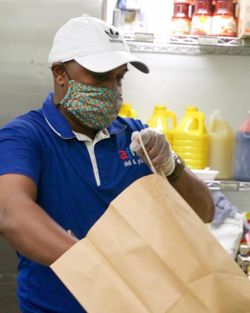 A man in a blue shirt and mask prepares a takeout bag in a kitchen filled with condiments and supplies.