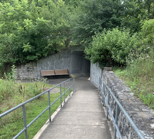 A paved path leads to a tunnel entrance, flanked by greenery and a bench on the side.