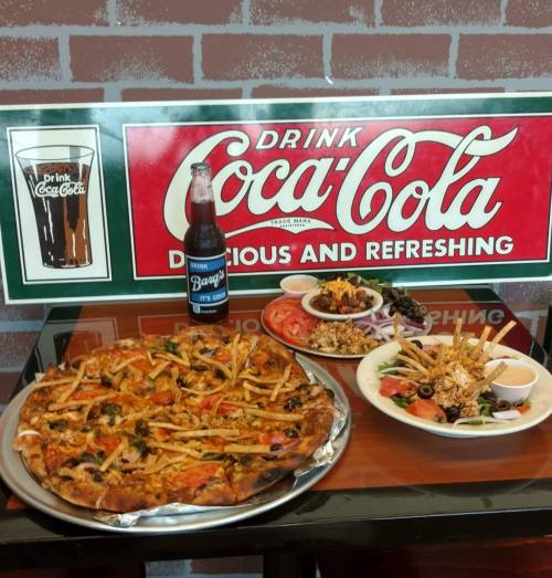 A table with a large pizza, a plate of salad, and a bottle of soda, set against a vintage Coca-Cola sign.